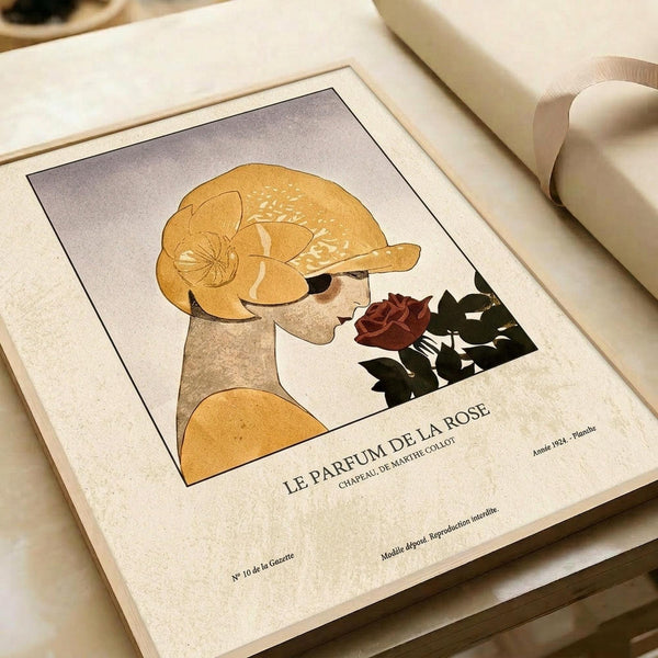 May include: Vintage-style art print featuring stylized woman in golden yellow hat smelling deep red rose, displayed in light wooden frame on cream textured paper, shown on neutral surface with rolled papers nearby in soft natural lighting