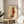 May include: Framed Paul Klee abstract art print displayed on light wood round side table next to beige upholstered chair with white pillow. Poster features geometric shapes in warm orange, rust, and teal tones with linear elements on cream background. Natural sunlight creates soft shadows on white wall, with green plant leaves visible in corner.