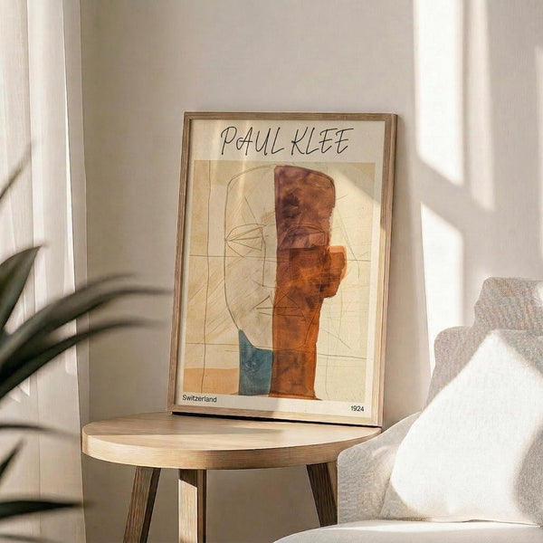 May include: Framed Paul Klee abstract art print displayed on light wood round side table next to beige upholstered chair with white pillow. Poster features geometric shapes in warm orange, rust, and teal tones with linear elements on cream background. Natural sunlight creates soft shadows on white wall, with green plant leaves visible in corner.
