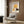 May include: Vintage-style art poster featuring woman in golden yellow hat holding dark red rose, mounted in light wood frame on warm brown wood paneled wall. Modern beige armchair with geometric patterned pillow sits below artwork. Golden floor lamp and dark round side table with open book complete the cozy reading nook scene with natural lighting from nearby window.