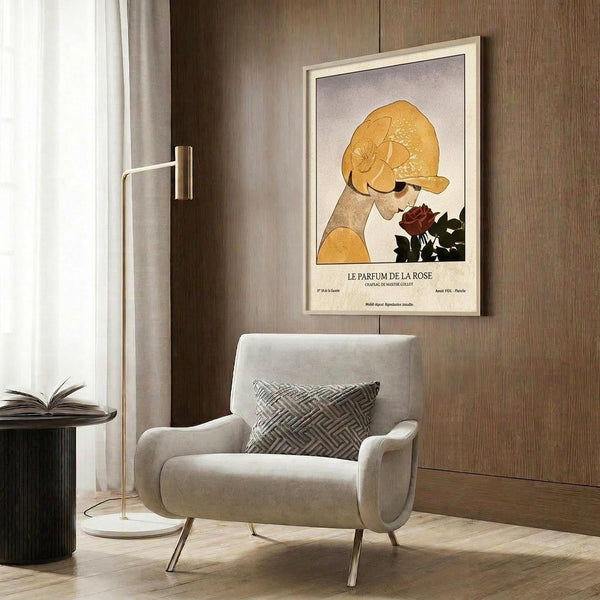 May include: Vintage-style art poster featuring woman in golden yellow hat holding dark red rose, mounted in light wood frame on warm brown wood paneled wall. Modern beige armchair with geometric patterned pillow sits below artwork. Golden floor lamp and dark round side table with open book complete the cozy reading nook scene with natural lighting from nearby window.