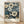 May include: Framed botanical art print featuring densely packed flowers in cream, white, and blue tones against a dark navy background. The artwork shows daisy-like blooms with detailed centers in a vintage illustrative style. Below the floral design is text reading "YAYOI KUSAMA" and Japanese characters, with "TOKYO" in the corner. The print sits in a light wood frame on wooden flooring against a white textured wall with soft natural lighting.