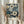 May include: Hands holding a framed botanical art print featuring white and blue daisy-like flowers on a dark navy background. The print has text at the bottom reading "YAYOI KUSAMA" and Japanese characters with "TOKYO". Person wears a cream-colored long-sleeved top with gathered cuffs and holds dried wheat stalks. Natural window light creates soft shadows on a neutral wall background.