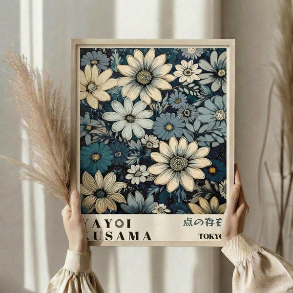 May include: Hands holding a framed botanical art print featuring white and blue daisy-like flowers on a dark navy background. The print has text at the bottom reading "YAYOI KUSAMA" and Japanese characters with "TOKYO". Person wears a cream-colored long-sleeved top with gathered cuffs and holds dried wheat stalks. Natural window light creates soft shadows on a neutral wall background.