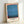 May include: Framed poster featuring abstract color field artwork with horizontal bands of blue and teal dominating the composition, accented by coral orange at the top and red elements along the edges. Text at bottom reads "THE MODERNIST ARCHIVE MARK ROTHKO HISTORY OF PAINTING CHROMA STUDIES 1950s MODERN ART". Light wood frame leaning against white wall corner, showing texture and grain of paper print.