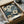 May include: Floral artwork displayed in black frame inside cardboard shipping box with tissue paper packaging. Design features cream and blue flowers with dark navy background in vintage botanical style. Box sits on white fabric surface next to wooden rolling pin and glass jar, suggesting home decor unboxing scene.