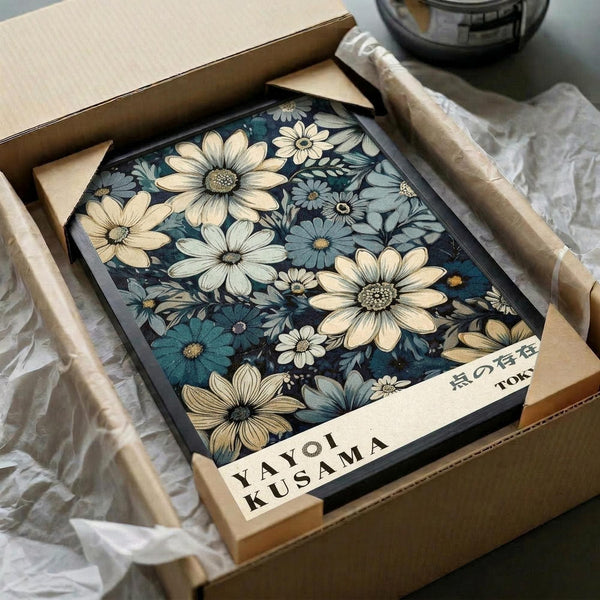 May include: Floral artwork displayed in black frame inside cardboard shipping box with tissue paper packaging. Design features cream and blue flowers with dark navy background in vintage botanical style. Box sits on white fabric surface next to wooden rolling pin and glass jar, suggesting home decor unboxing scene.