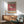 May include: Framed exhibition poster featuring a detailed pufferfish illustration on pink background with black text and Japanese characters, displayed above mid-century modern wooden credenza with white ceramic lamp, alongside sage green upholstered armchair with wooden frame in sunlit room with white walls.