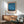 May include: Framed Mark Rothko exhibition poster featuring abstract blue and orange color field painting displayed on white wall above mid-century modern wooden credenza. Green upholstered chair with wooden arms sits nearby, white ceramic table lamp on credenza surface. Soft natural lighting creates shadows across the minimalist interior scene.