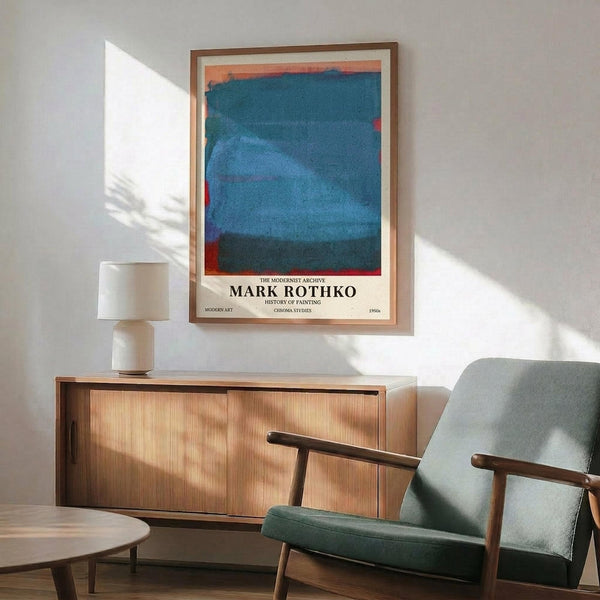 May include: Framed Mark Rothko exhibition poster featuring abstract blue and orange color field painting displayed on white wall above mid-century modern wooden credenza. Green upholstered chair with wooden arms sits nearby, white ceramic table lamp on credenza surface. Soft natural lighting creates shadows across the minimalist interior scene.