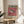 May include: Framed exhibition poster featuring a detailed pufferfish illustration on pink background, displayed on wooden chair beside neutral couch. Natural lighting creates soft shadows on white wall. Poster shows vintage gallery exhibition details from San Francisco Japantown, with Japanese text at bottom. Green plant leaves visible in corner, creating minimalist modern home decor scene.