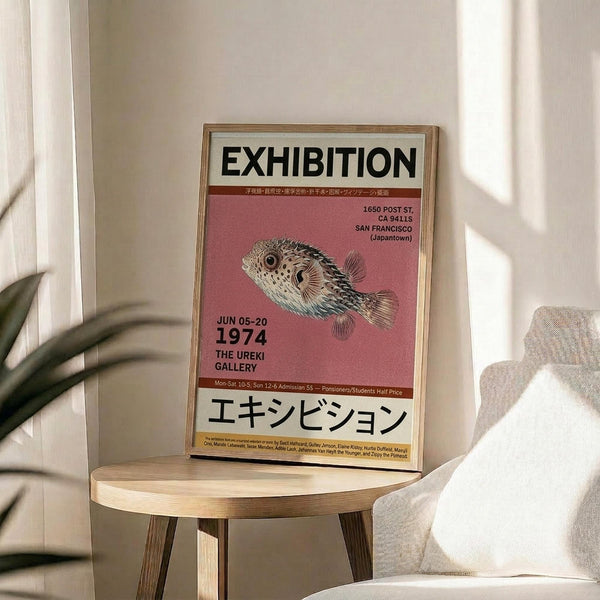 May include: Framed exhibition poster featuring a detailed pufferfish illustration on pink background, displayed on wooden chair beside neutral couch. Natural lighting creates soft shadows on white wall. Poster shows vintage gallery exhibition details from San Francisco Japantown, with Japanese text at bottom. Green plant leaves visible in corner, creating minimalist modern home decor scene.