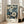 May include: Framed botanical art print leaning against white wall next to beige sofa in bright living room. Artwork features densely packed flowers in cream, blue, and teal tones on dark background with "YAYOI KUSAMA" and Japanese text below. Natural wood frame, multiple windows with sunlight streaming in, hardwood floors visible.