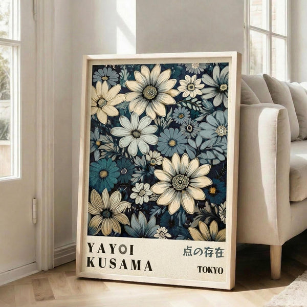 May include: Framed botanical art print leaning against white wall next to beige sofa in bright living room. Artwork features densely packed flowers in cream, blue, and teal tones on dark background with "YAYOI KUSAMA" and Japanese text below. Natural wood frame, multiple windows with sunlight streaming in, hardwood floors visible.
