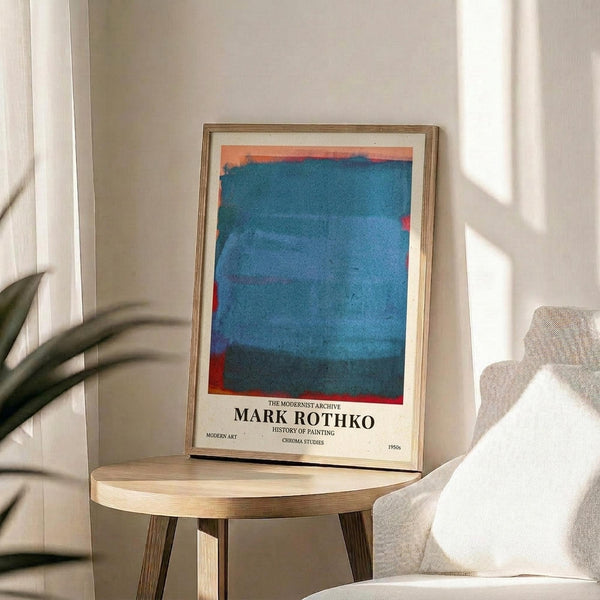 May include: Framed poster featuring Mark Rothko's abstract color field painting with vibrant blue brushstrokes and orange-red accents at top, displayed on light wood round side table next to beige upholstered chair. Natural sunlight streams across white walls creating geometric shadows, with green plant leaves visible in corner of minimalist modern living room setting.