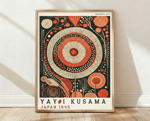 May include: Framed modern art poster with large concentric circles in red, cream, and black, surrounded by abstract botanical patterns and geometric shapes. The artwork features intricate line work and dotted details in a folk art style. Text reads "Yayoi Kusama Japan 1945" below the main design. Light wood frame sits on wooden flooring against white wall with soft natural lighting.