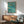 May include: Framed poster with teal watercolor background showing three orange swimmers diving through stylized waves, with white and Japanese text at top. Displayed above wooden sideboard with white lamp in modern living room setting with mint green upholstered chair and round coffee table. Warm natural lighting creates geometric shadows on white wall.