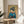 May include: Framed Japanese-style artwork showing a geisha with elaborate blue-black hair adorned with gold hair ornaments, wearing a blue kimono with red collar and floral obi sash. She holds thin sticks while surrounded by cherry blossom branches on a golden yellow background. The poster sits on a light wood round side table next to a beige upholstered chair in a bright room with white curtains and plant shadows.