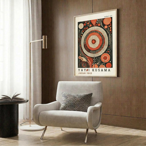 May include: Framed abstract art print with large central circle surrounded by smaller circular and geometric patterns in red, orange, black and cream colors, displayed on wood-paneled wall above beige upholstered chair with geometric throw pillow, brass floor lamp and dark round side table with open book visible in modern interior setting.