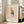 May include: Framed art print leaning against white wall next to beige upholstered chair in bright living room. Poster features geometric owl design in red, yellow, blue and orange angular shapes with thin black lines for legs. Text reads "PICASSO" at top and "The Met L'Art Minimaliste" at bottom on cream background. Natural wood frame, sunlight streaming through windows onto hardwood floors.