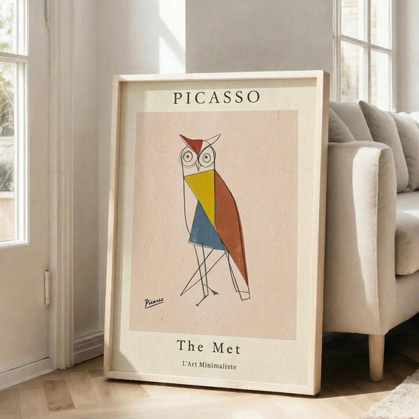 May include: Framed art print leaning against white wall next to beige upholstered chair in bright living room. Poster features geometric owl design in red, yellow, blue and orange angular shapes with thin black lines for legs. Text reads "PICASSO" at top and "The Met L'Art Minimaliste" at bottom on cream background. Natural wood frame, sunlight streaming through windows onto hardwood floors.
