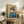 May include: Framed Japanese-style artwork showing a geisha in blue kimono with elaborate dark hair ornament, set against golden background with cherry blossoms, displayed on rustic wooden sideboard alongside books, ceramic vase, and fern plant in modern living room with beige chair and neutral walls.