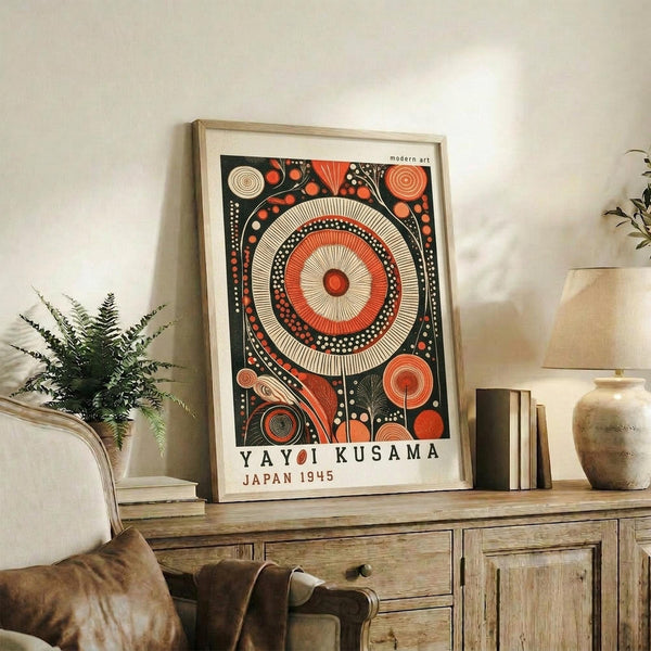 May include: Framed vintage-style poster with large central orange and cream spiral surrounded by smaller circular patterns on dark background, displayed on rustic wooden sideboard beside fern plant, ceramic vase, and stacked books in neutral-toned living room setting
