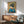 May include: Framed Japanese-style artwork showing a geisha in blue kimono with elaborate dark hair and hair ornaments, set against golden background with cherry blossoms. Displayed above mid-century modern wooden credenza with table lamp, next to sage green upholstered armchair in bright, naturally-lit room with white walls.