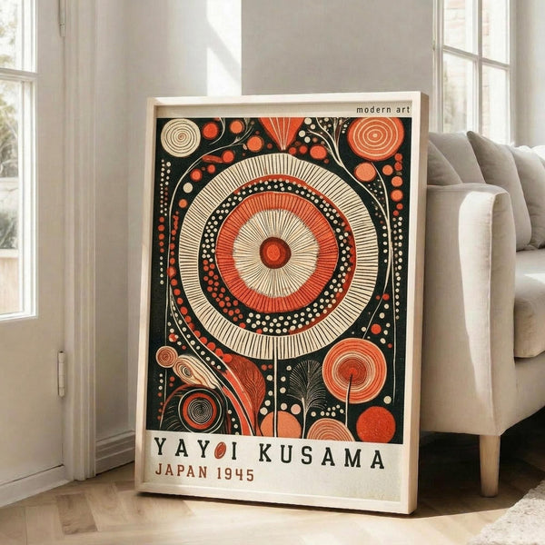 May include: A framed modern art print featuring a large circular mandala design with concentric rings in orange, cream, and black colors, surrounded by decorative dots and spiral patterns. The poster has "YAYOI KUSAMA JAPAN 1945" text at the bottom and displays in a light wood frame, leaning against a white wall next to a beige sofa in a bright living room with large windows.