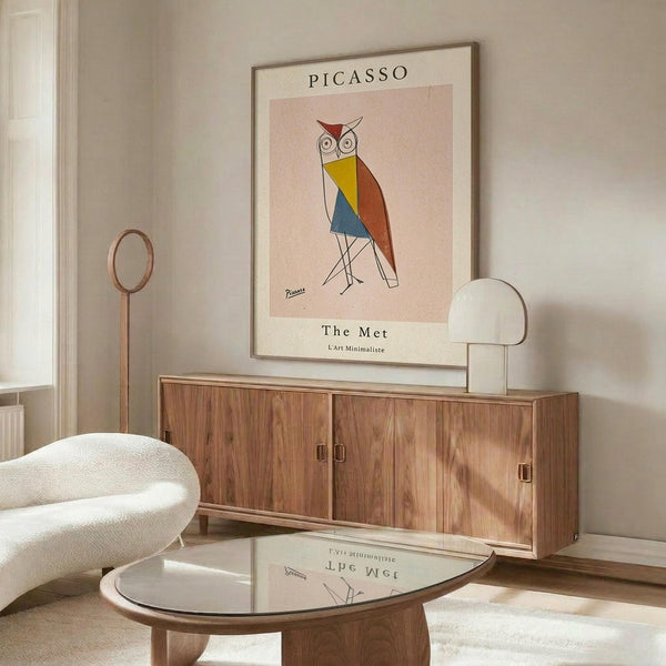 May include: Framed art print featuring a geometric owl in Picasso's style with colorful angular shapes in red, yellow, blue and orange against a pink background. Print hangs above a walnut wood credenza in a minimalist room with white upholstered chair, glass coffee table, and modern lamp. Warm natural lighting creates subtle shadows across the neutral-toned space.