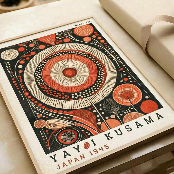 Abstract art poster featuring a large central mandala with concentric circles in orange, cream, and black. The design includes radiating lines, dotted patterns, and smaller circular elements scattered around the composition. The artwork has "modern art" at the top and "YAYOI KUSAMA JAPAN 1945" at the bottom. The print is shown flat on a cream surface next to rolled paper tubes, displaying the geometric pattern's intricate details and bold color contrast.