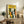 May include: Framed poster with yellow background featuring illustrated cats in various colors including black, orange, striped, and white with "FELINES" text at top. Wood frame sits on rustic wooden sideboard next to fern plant, books, ceramic vase, and lamp in neutral-toned living room setting with beige chair visible.