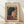 May include: Framed poster in light wood frame featuring stylized portrait of woman with dark bob haircut in profile, surrounded by colorful flowers and leaves in red, blue, and green on cream background. Frame leans against white wall on light wooden floor with natural lighting casting shadows.