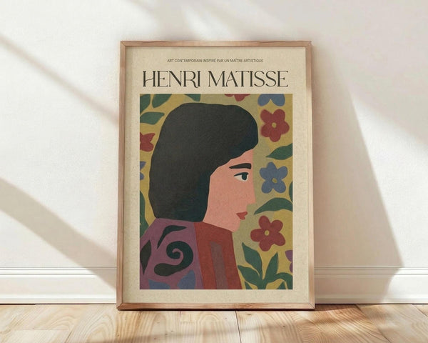 May include: Framed poster in light wood frame featuring stylized portrait of woman with dark bob haircut in profile, surrounded by colorful flowers and leaves in red, blue, and green on cream background. Frame leans against white wall on light wooden floor with natural lighting casting shadows.