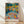 May include: Framed art print leaning against white wall on light wood floor with natural window light creating shadows. The colorful artwork depicts people gathered by water surrounded by vibrant trees in blues, oranges, yellows and greens in an impressionist style. Text reads "HENRI MATISSE PARIS" below the image. Natural wood frame visible around the poster.
