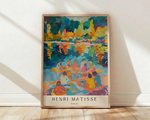 May include: Framed art print leaning against white wall on light wood floor with natural window light creating shadows. The colorful artwork depicts people gathered by water surrounded by vibrant trees in blues, oranges, yellows and greens in an impressionist style. Text reads "HENRI MATISSE PARIS" below the image. Natural wood frame visible around the poster.