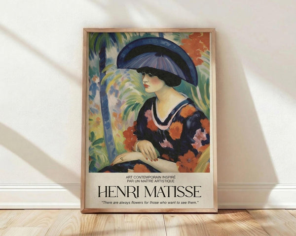 May include: Framed art print featuring a woman wearing a large blue and black hat with floral dress in vibrant colors including orange, pink, and navy. The painting shows tropical foliage in the background with palm fronds. Below the artwork is text reading "HENRI MATISSE" and a quote about flowers. The poster has a light wooden frame and sits on wooden flooring against a white wall with natural lighting creating shadows.