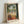 May include: Framed art print in light wood frame leaning against white wall. Tropical scene shows figure in orange dress on terrace overlooking water, with palm trees, ornate archway, and fruit bowl on small table. Warm colors include greens, oranges, and blues in painterly style reminiscent of mid-century modern art.