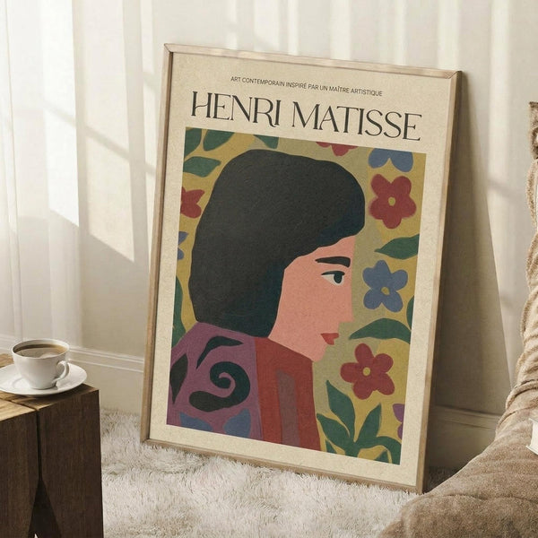 May include: Framed art print featuring a woman with dark hair in profile against a colorful floral background with red, blue and green flowers on yellow. The poster has "HENRI MATISSE" text at top and shows contemporary art styling. Displayed leaning against white wall near wooden stool with coffee cup, on textured white rug beside beige blanket.