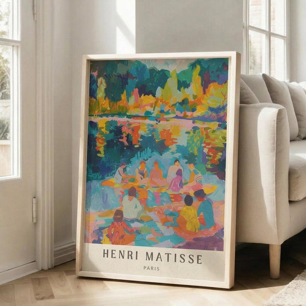 May include: Colorful Henri Matisse art print in light wooden frame leaning against white wall next to beige sofa. The vibrant poster shows figures by water with lush foliage in blues, greens, oranges and yellows. Natural sunlight streams through tall windows onto hardwood floors in this bright, minimalist living room setting.