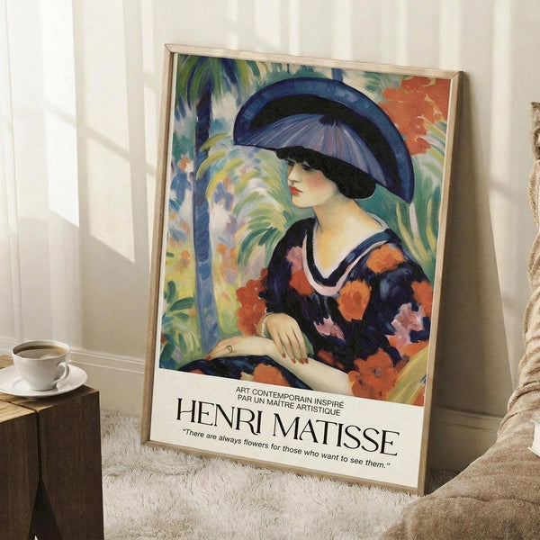 May include: Framed Henri Matisse exhibition poster featuring a woman in a large blue hat and colorful floral dress, painted in vibrant blues, oranges, and greens. The poster displays French text and a Matisse quote about flowers. Shown leaning against a white wall in a bright room setting with natural light, positioned next to a coffee cup on a wooden surface and a cream knitted throw blanket on a white textured rug.