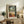 May include: Framed Henri Matisse-style poster showing tropical terrace scene with palm trees, figures in white and orange clothing overlooking water, fruit bowl on small table, displayed on rustic wooden console with fern plant, ceramic vase, and books in neutral-toned living room setting