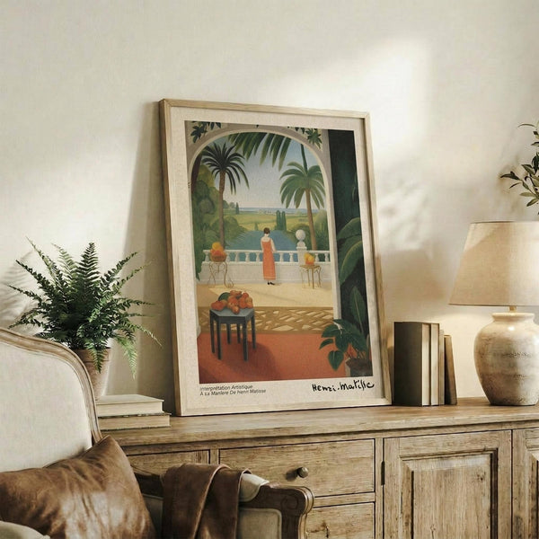 May include: Framed Henri Matisse-style poster showing tropical terrace scene with palm trees, figures in white and orange clothing overlooking water, fruit bowl on small table, displayed on rustic wooden console with fern plant, ceramic vase, and books in neutral-toned living room setting