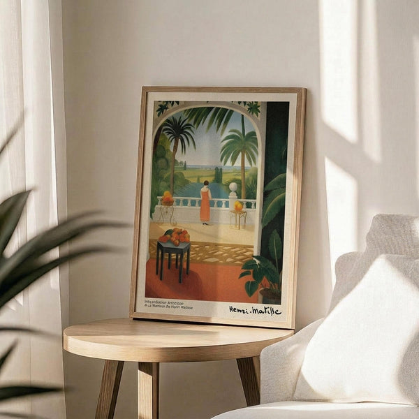 May include: Framed art print displayed on round wooden side table next to beige upholstered chair. The colorful artwork depicts a tropical terrace scene with palm trees, figures in flowing dresses on a balcony overlooking water, and a small table with fruit. Natural sunlight creates shadows across white walls, with a plant visible in the corner of the room.