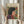 May include: Hands holding a framed art print featuring a stylized woman with dark bob haircut wearing a purple and coral garment, surrounded by colorful flowers on yellow background. Light wood frame, dried pampas grass visible to left, soft beige clothing and neutral wall backdrop create warm, minimalist styling.