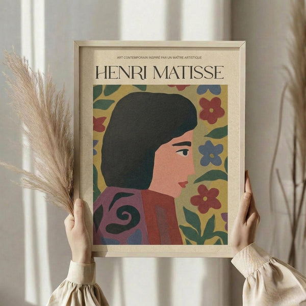 May include: Hands holding a framed art print featuring a stylized woman with dark bob haircut wearing a purple and coral garment, surrounded by colorful flowers on yellow background. Light wood frame, dried pampas grass visible to left, soft beige clothing and neutral wall backdrop create warm, minimalist styling.
