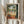 May include: Person in cream ruffled sleeves holding framed art print featuring Mediterranean terrace scene with woman in orange dress overlooking tropical landscape with palm trees, white balustrade, and fruit table. Dried pampas grass visible alongside neutral curtained background.