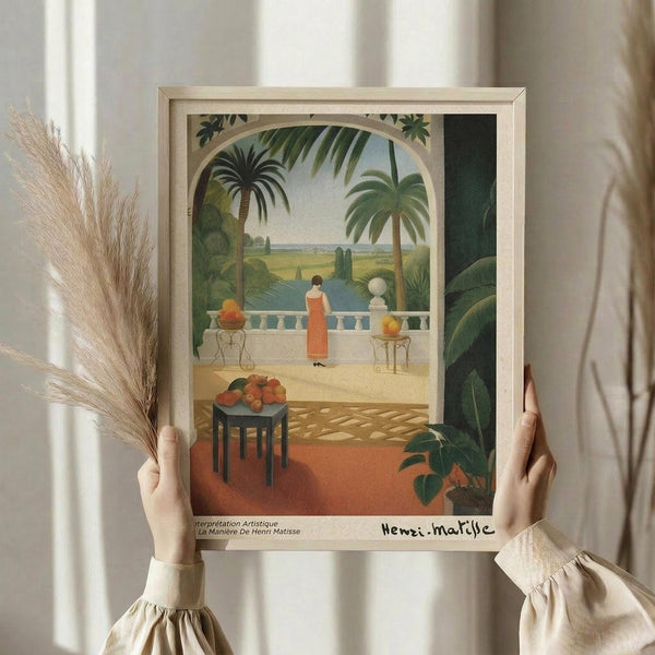 May include: Person in cream ruffled sleeves holding framed art print featuring Mediterranean terrace scene with woman in orange dress overlooking tropical landscape with palm trees, white balustrade, and fruit table. Dried pampas grass visible alongside neutral curtained background.