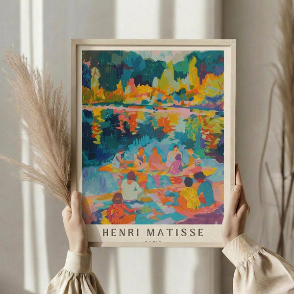 May include: Person in beige sweater holding framed colorful art print showing figures by water with vibrant trees. Fauvism style painting with bold blues, oranges, yellows, and greens depicting people gathered near a lake or river. Light wood frame with "HENRI MATISSE PARIS" text below artwork. Dried pampas grass and neutral curtains visible in bright, minimalist room setting.