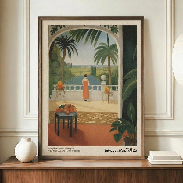 May include: Framed art print in warm tropical scene showing woman in coral dress on terrace overlooking palm trees and water. Matisse-inspired style with green foliage, archway, fruit bowl on dark table. Wood frame on credenza beside white ceramic vase and stacked books in neutral room with paneled walls.